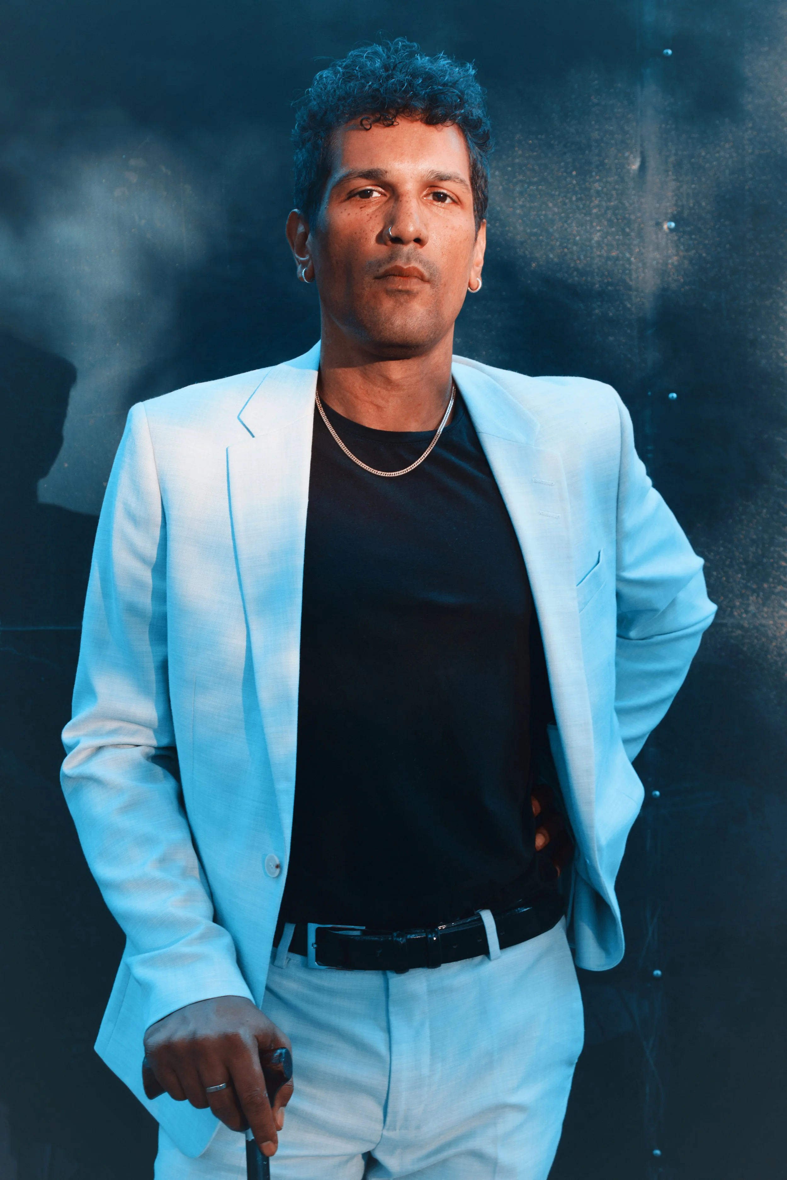 A person with dark, curly hair, wearing a white blazer, black shirt, and light-colored pants, standing against a dark background.