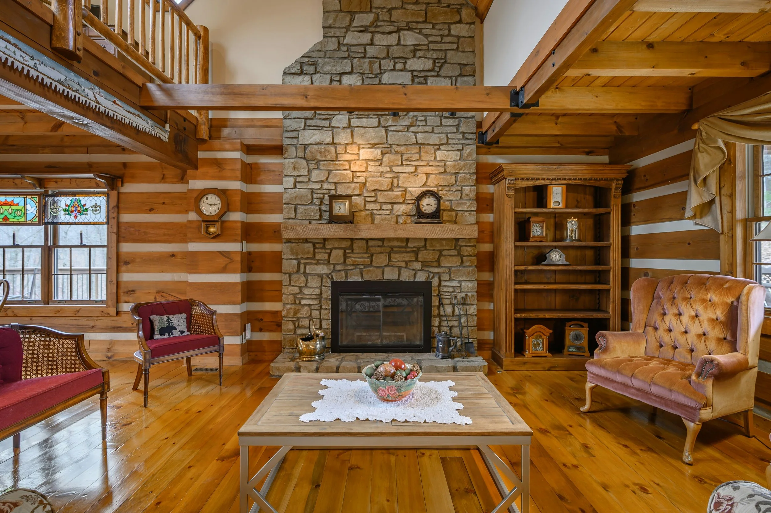Interior view of a rustic living room with a stone fireplace, wooden beams, wooden furniture, and stained glass windows.