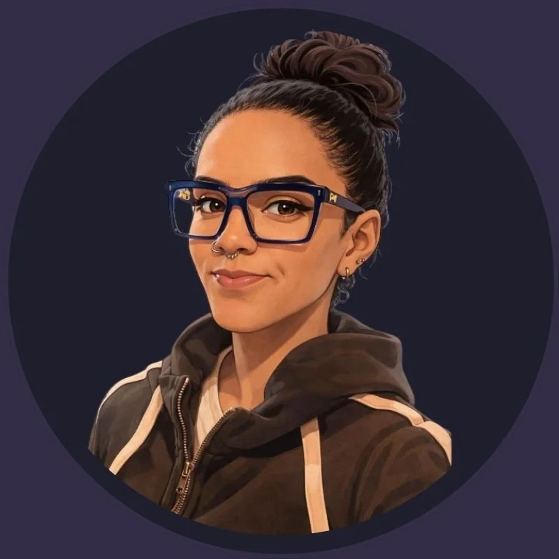 A young woman with glasses, a septum piercing, and earrings, smiling slightly, wearing a hoodie and standing against a dark background.