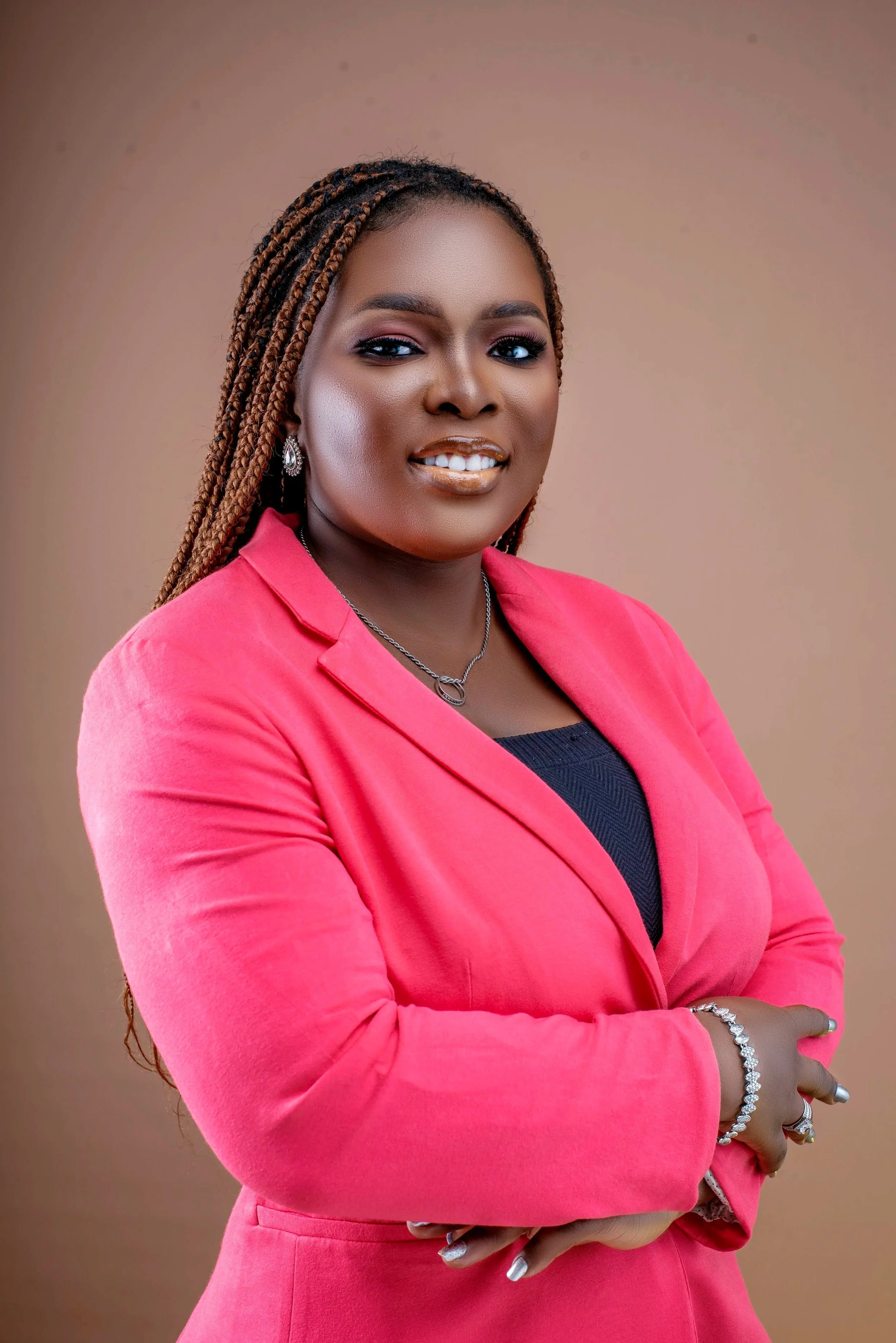 A woman with medium-dark skin in a pink blazer, wearing makeup, jewelry, and with styled hair, smiling with arms crossed, against a brown background.