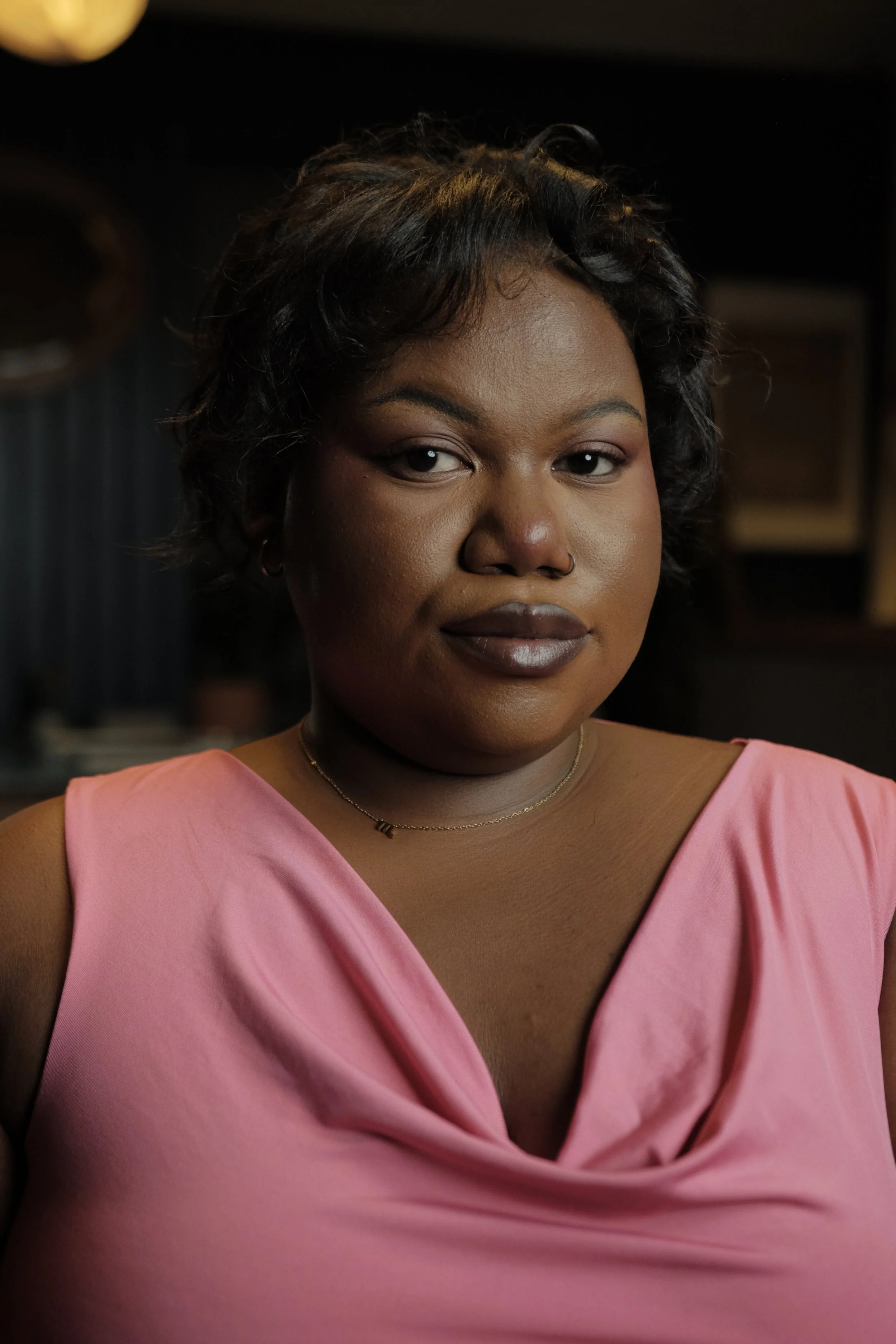 A woman with dark hair styled in soft curls, wearing a sleeveless pink top, a delicate gold necklace, and small hoop earrings, looking directly at the camera with a neutral expression in a dimly lit environment.
