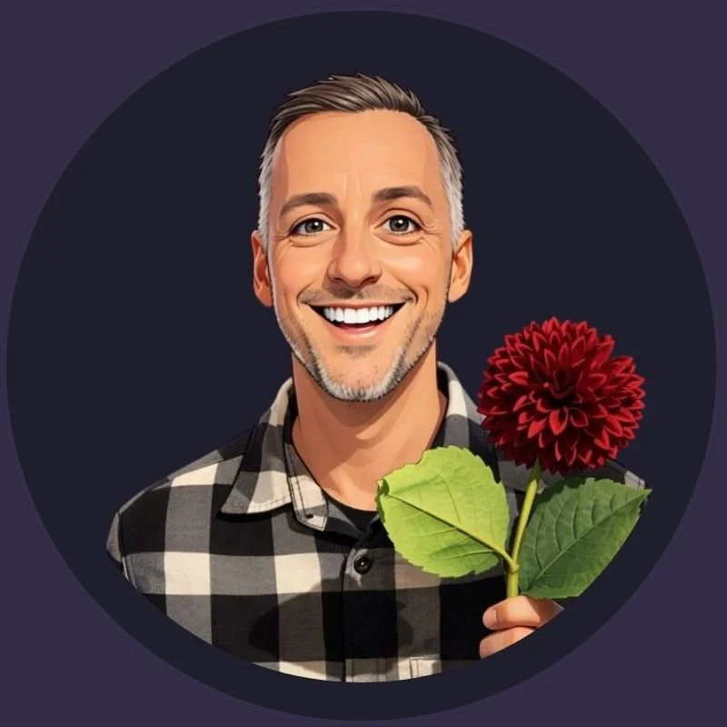 A smiling man with short gray hair, a beard, and wearing a black and white checkered shirt, holding a red dahlia flower and green leaves.