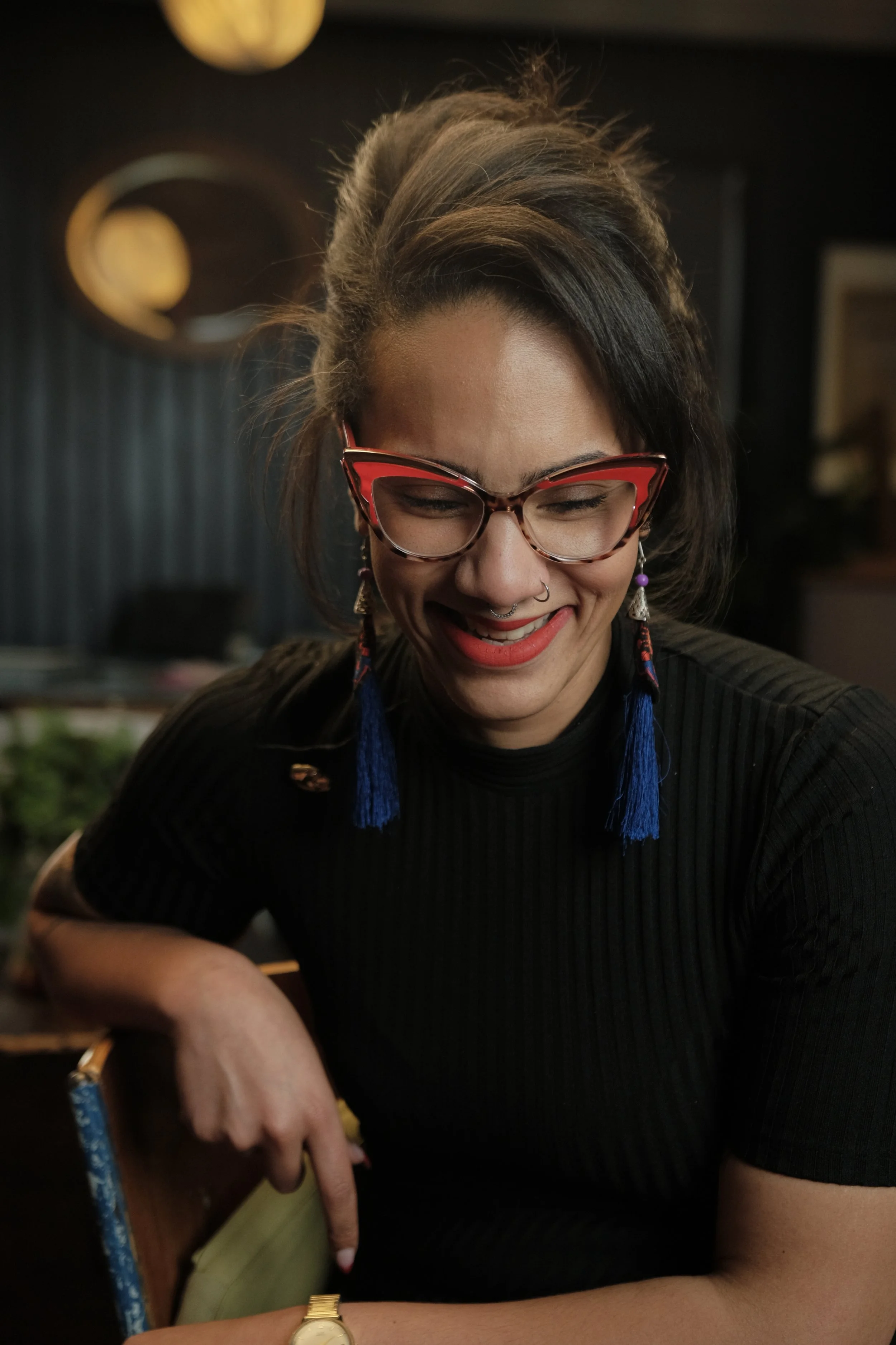 A woman with dark hair, wearing red cat-eye glasses, laughing with her eyes closed, dressed in black, with blue tassel earrings and a gold watch, sitting in a dimly lit room.