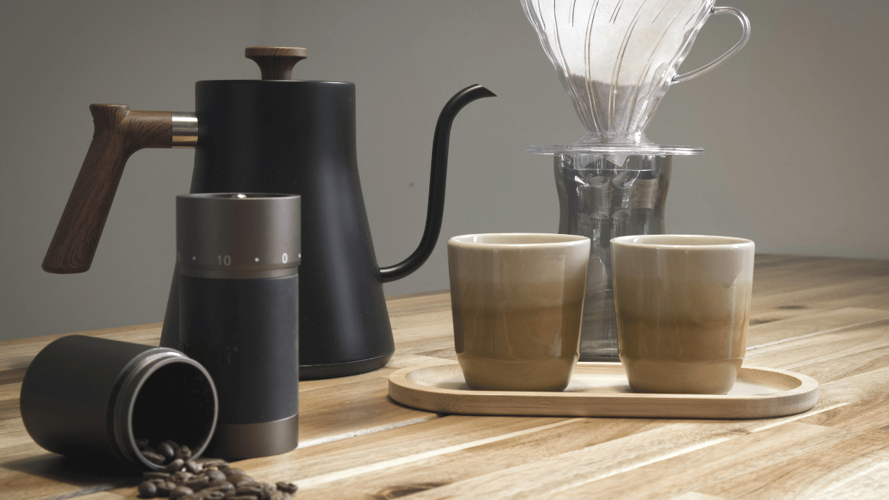 A coffee brewing setup on a wooden table featuring an electric coffee grinder, a pour-over coffee maker, two beige ceramic cups on a wooden tray, and a black stovetop kettle with a wooden handle.