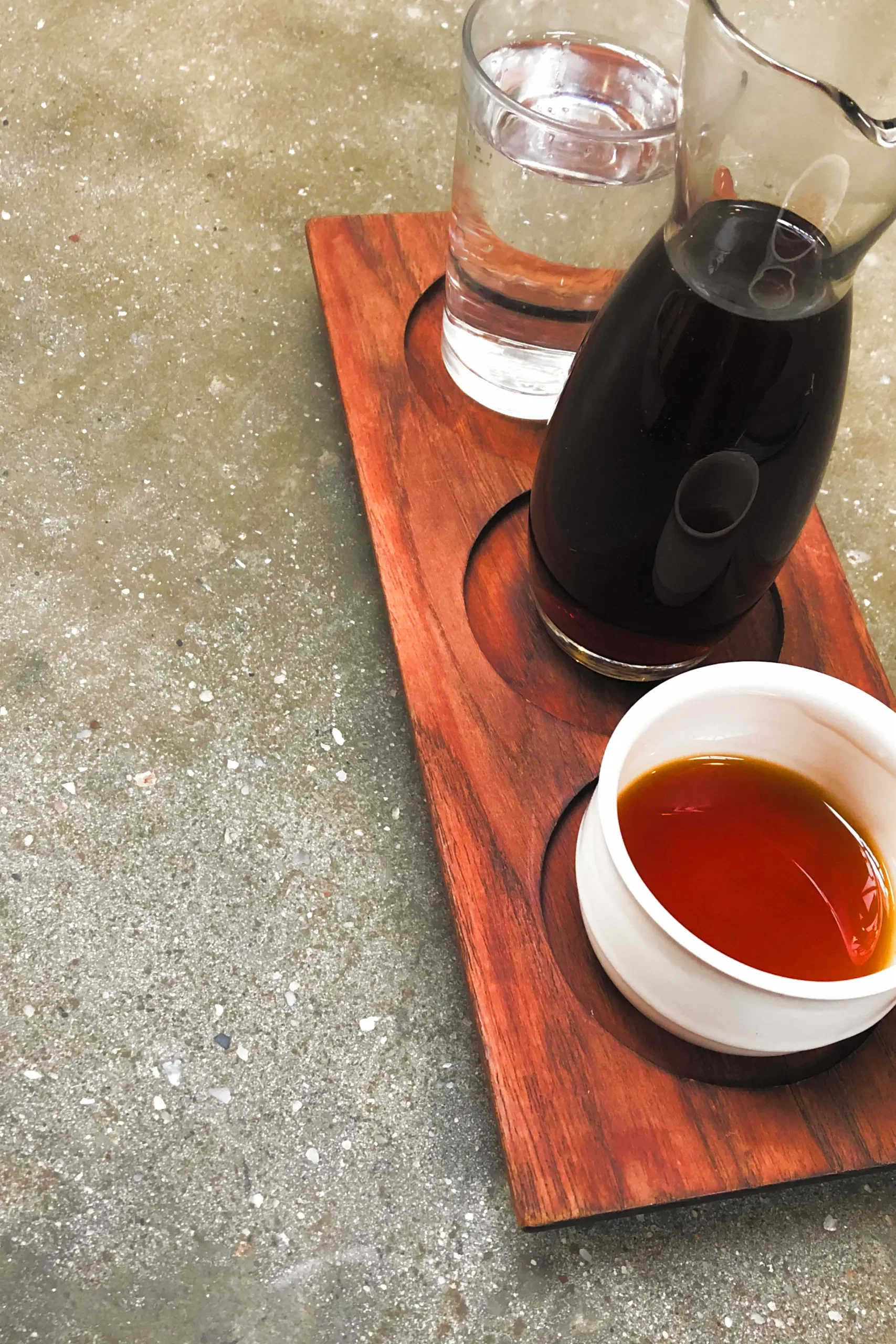 A wooden tray on a concrete surface holding a carafe of coffee, a glass of water, and a wide white bowl-shaped cup.
