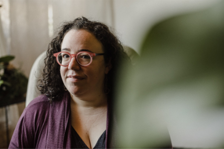 A woman with curly hair wearing red glasses, sitting indoors with a blurred green plant in the foreground.