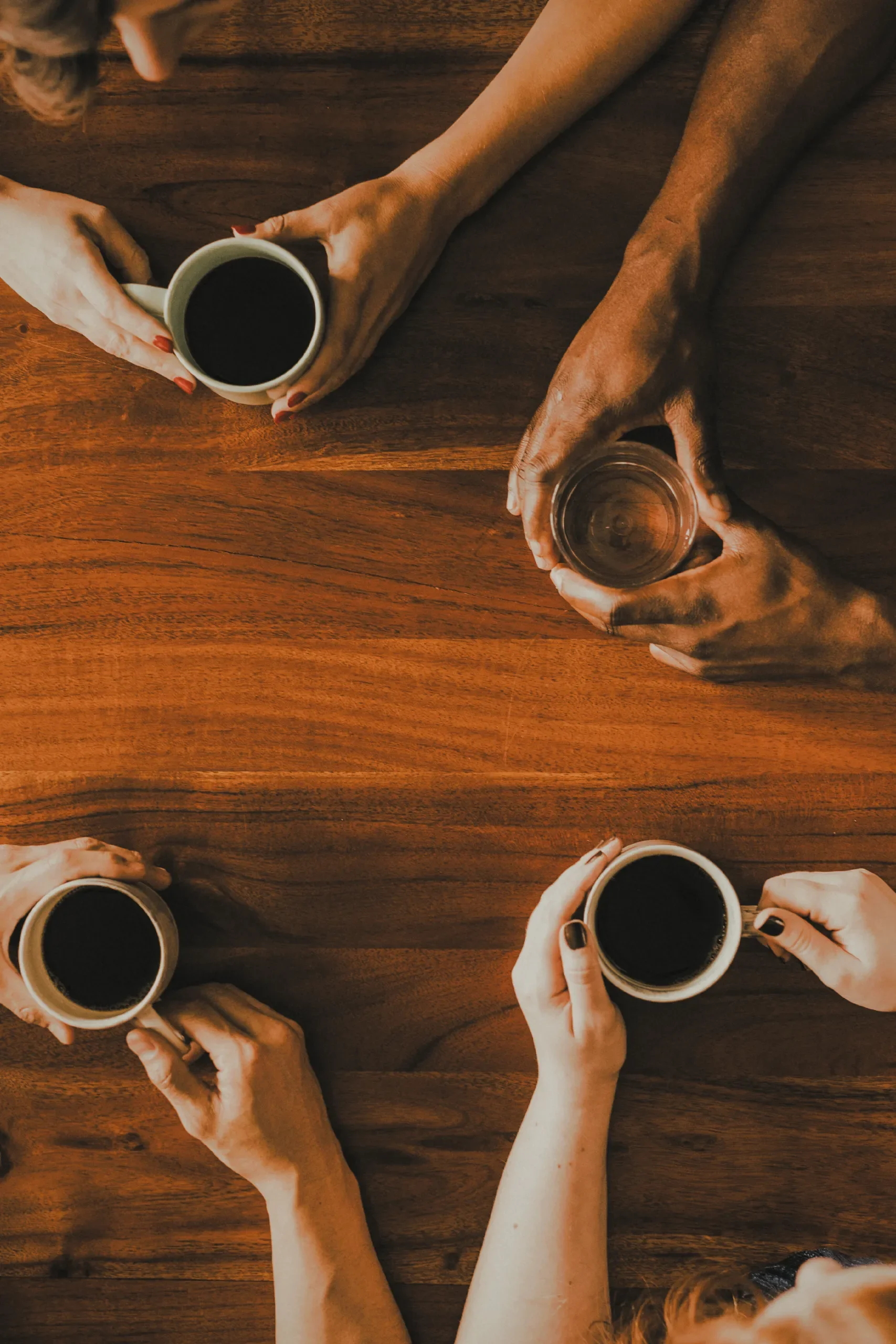 Four people sitting around a wooden table each holding a cup of coffee, with one person also holding a glass of water, viewed from above.
