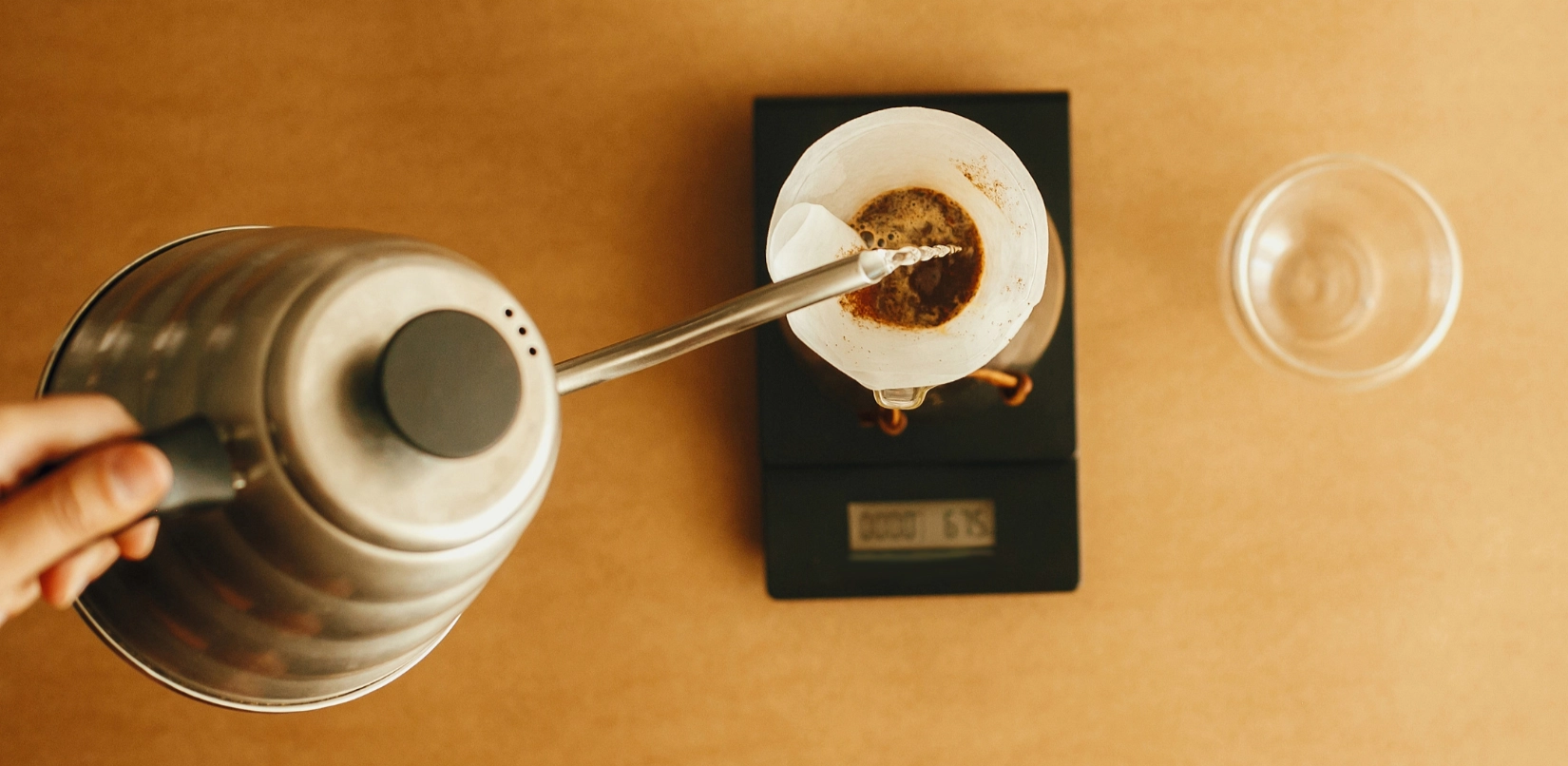 Top view of a hand holding an electric kettle pouring hot water into a coffee filter on a scale, with a glass of water to the right on a beige surface.