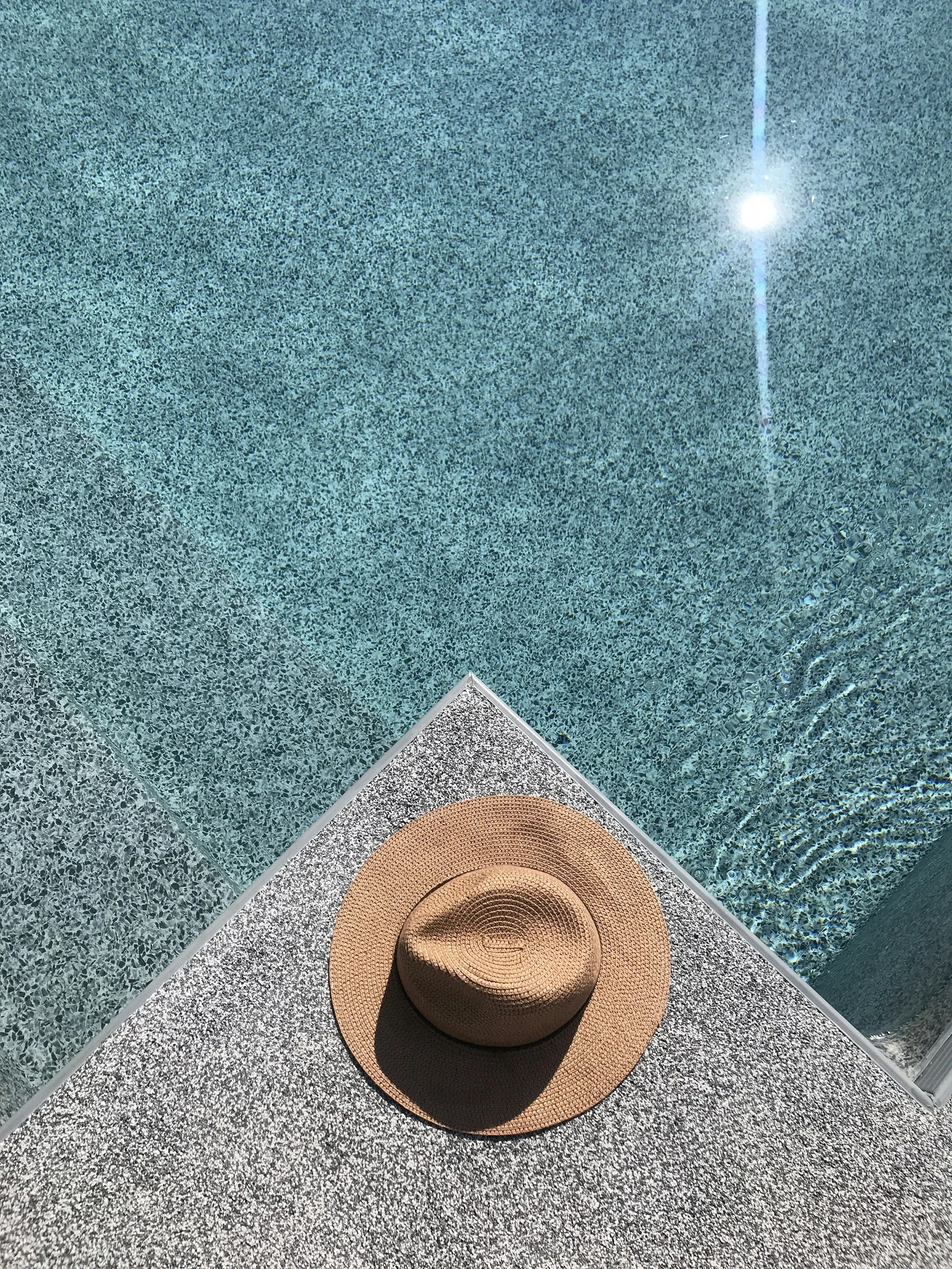 A woven sun hat with a wide brim resting on a textured surface, beside a swimming pool with clear blue water and a pool edge.