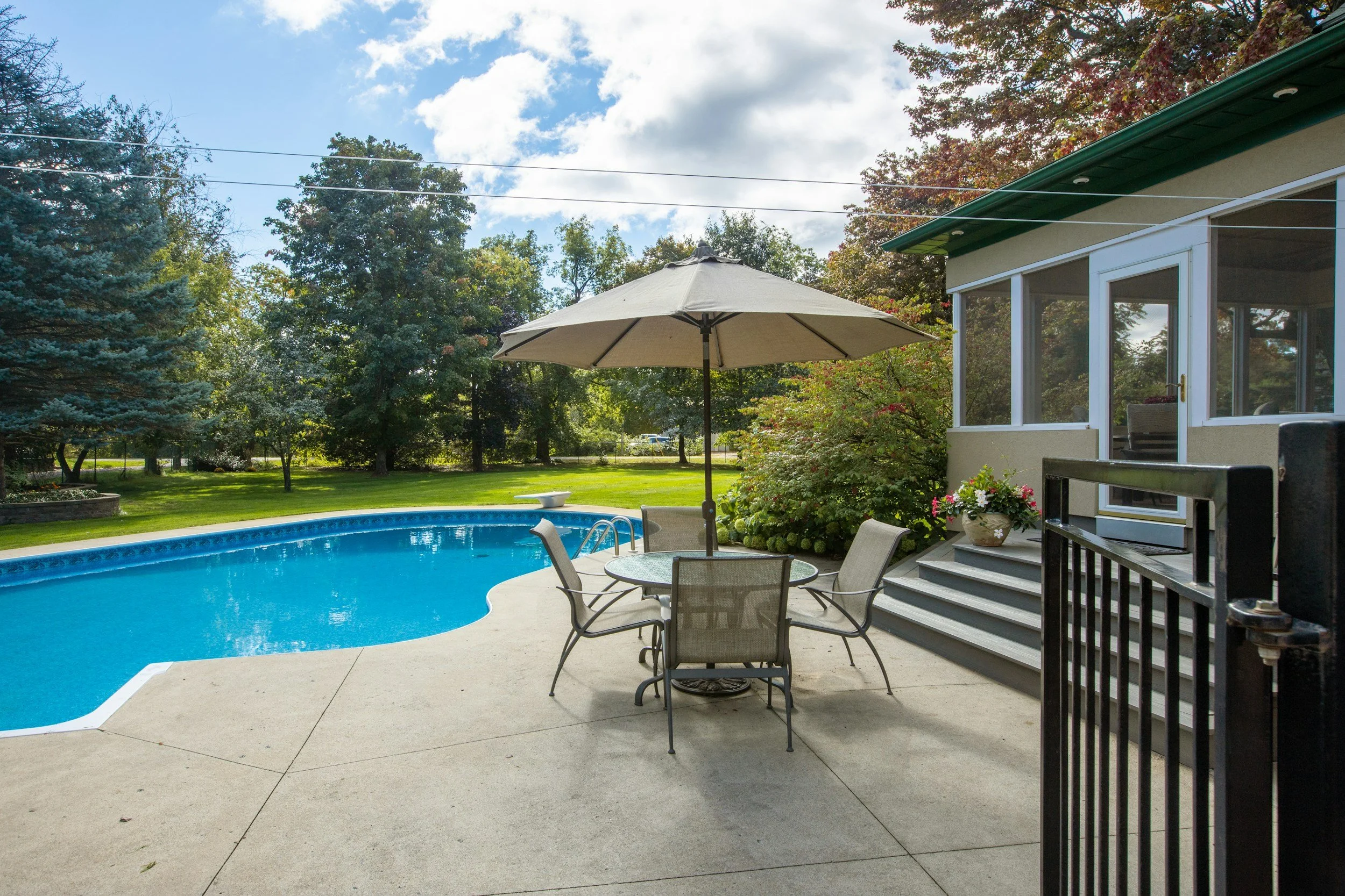 View of a backyard swimming pool with a patio table and chairs, a large umbrella, surrounded by greenery and trees, with a house or enclosed porch nearby.