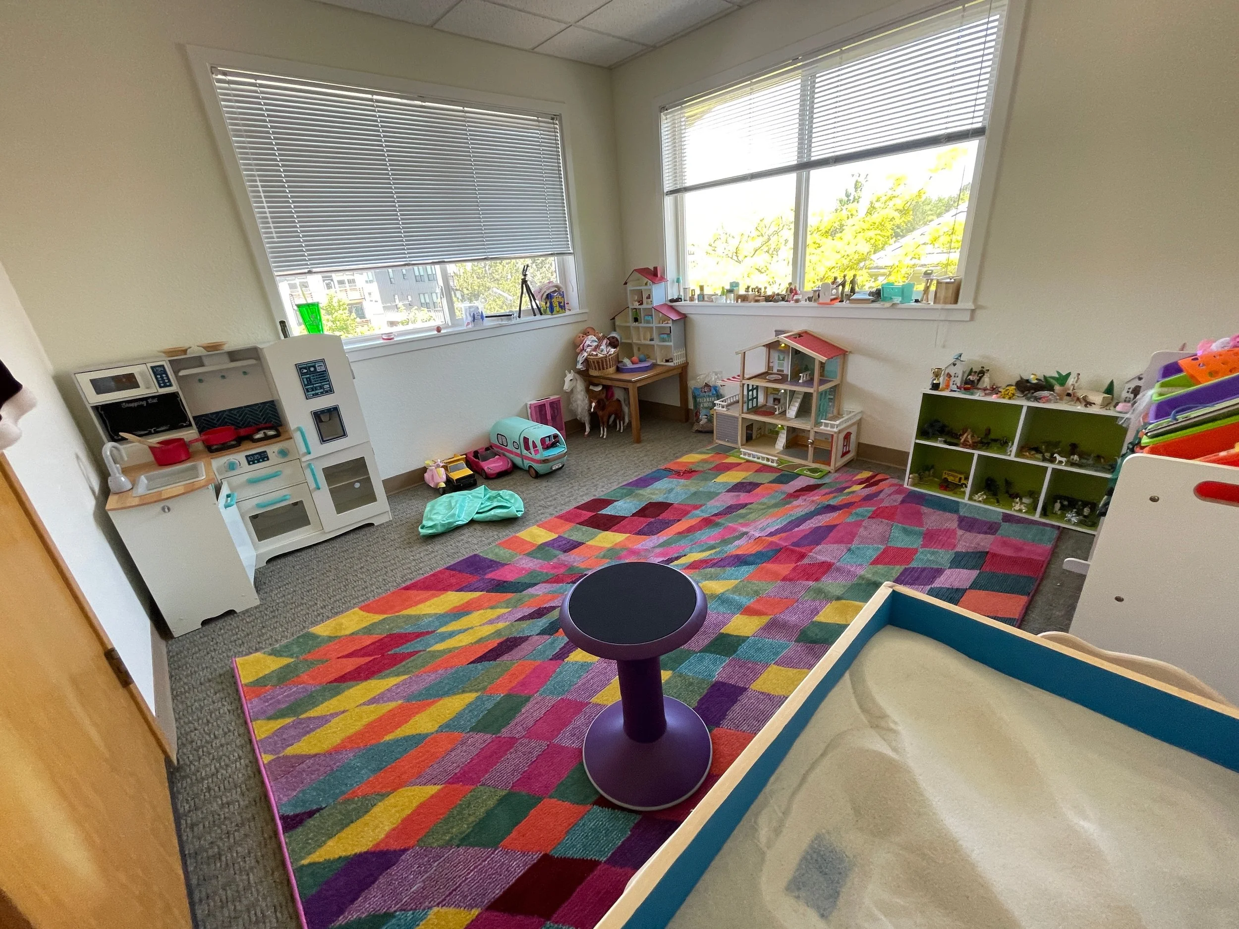 A colorful children's playroom with a multicolored geometric rug, toy kitchen set, small dollhouse, and various toys on shelves and windowsills, illuminated by large windows.