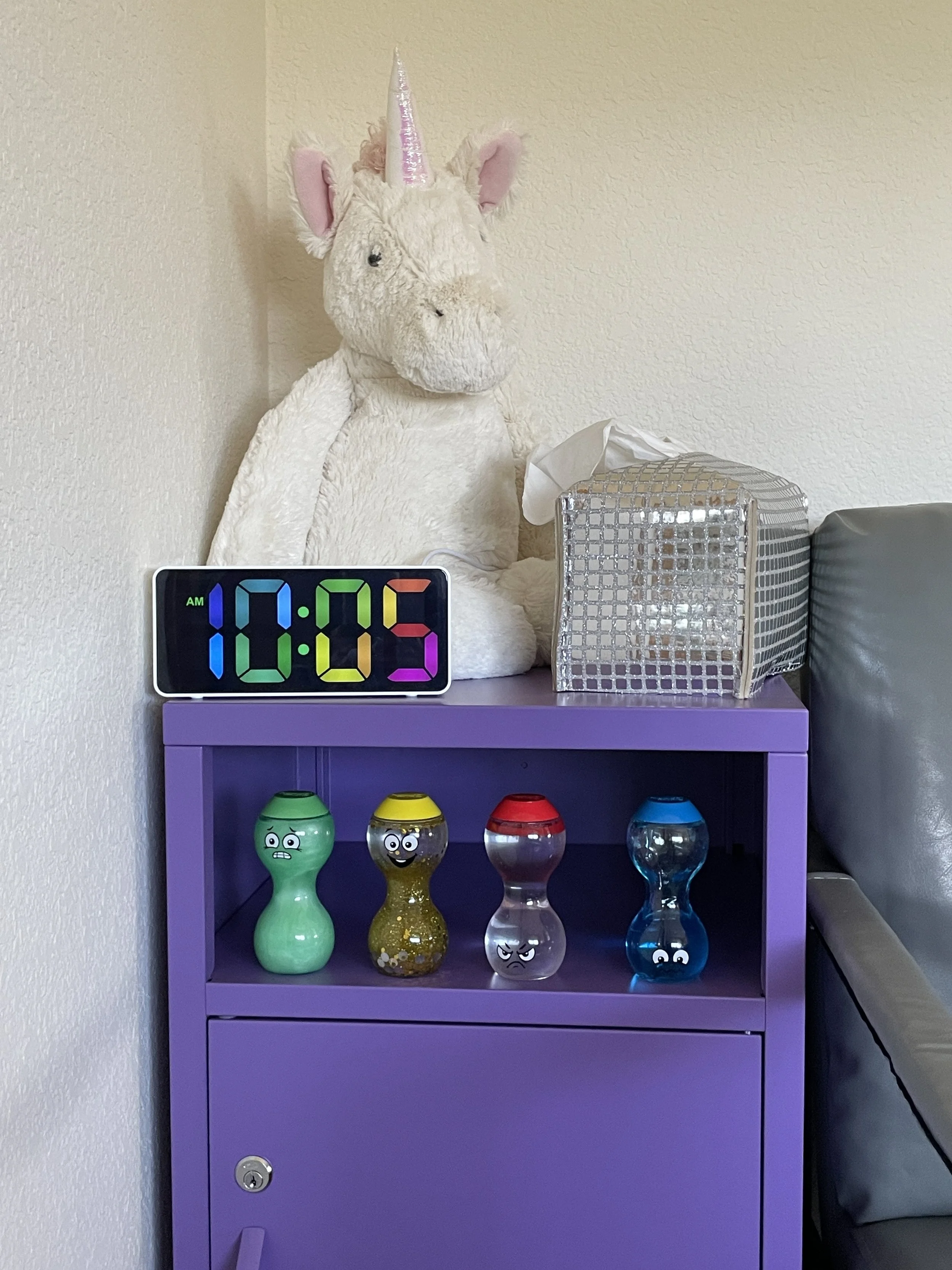 A purple cabinet with a plush unicorn toy on top, a digital clock displaying 10:05 AM, a tissue box, and four colorful finger skateboards with cartoon faces on the shelf.