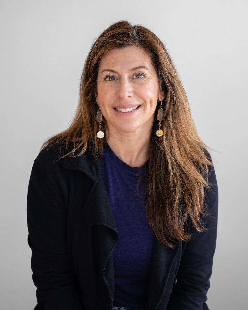 A woman with long brown hair, wearing a black jacket and blue top, smiling with a plain light gray background.