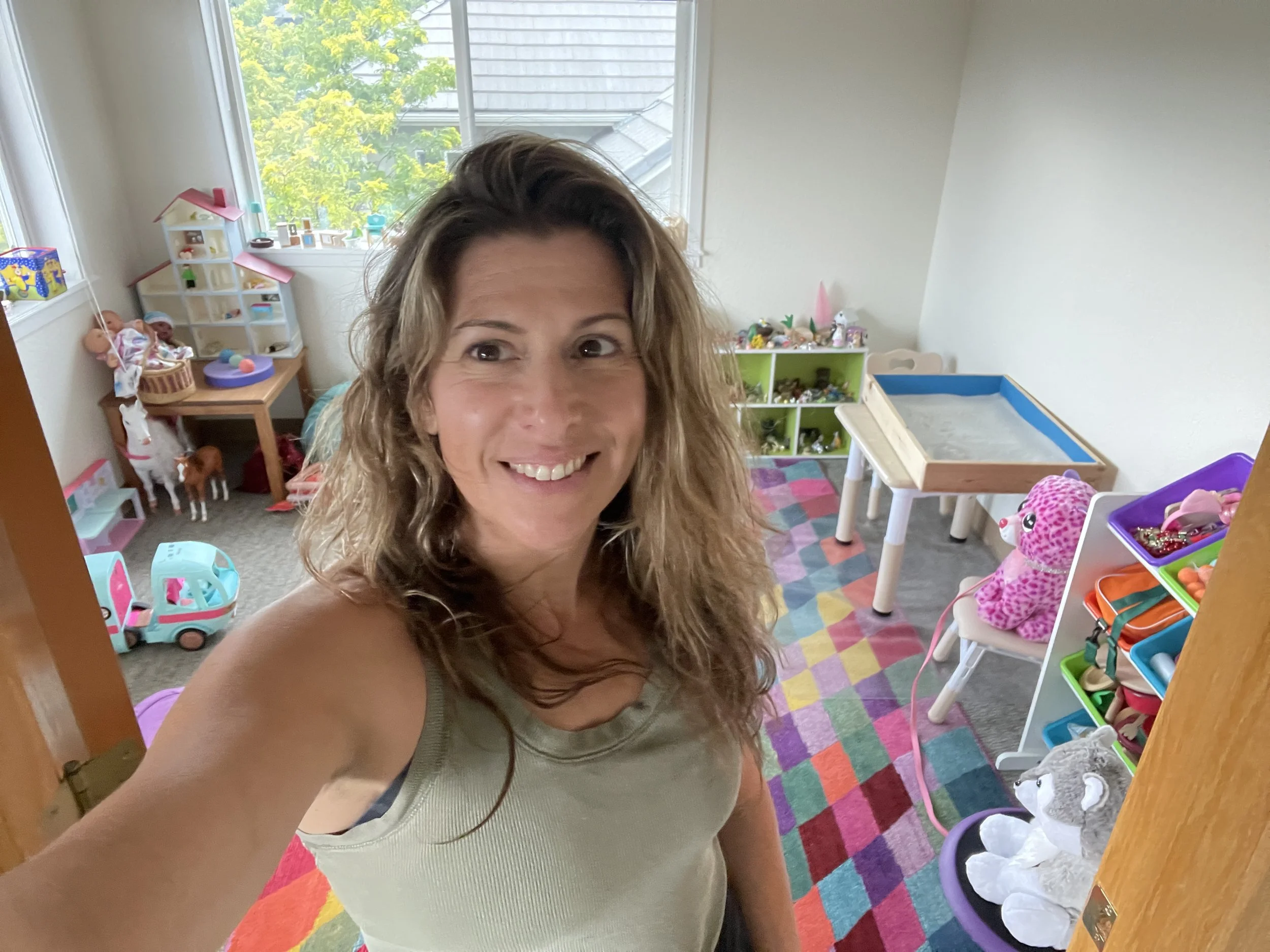 Woman taking a selfie in a colorful playroom filled with toys, including dolls, stuffed animals, and toy vehicles.