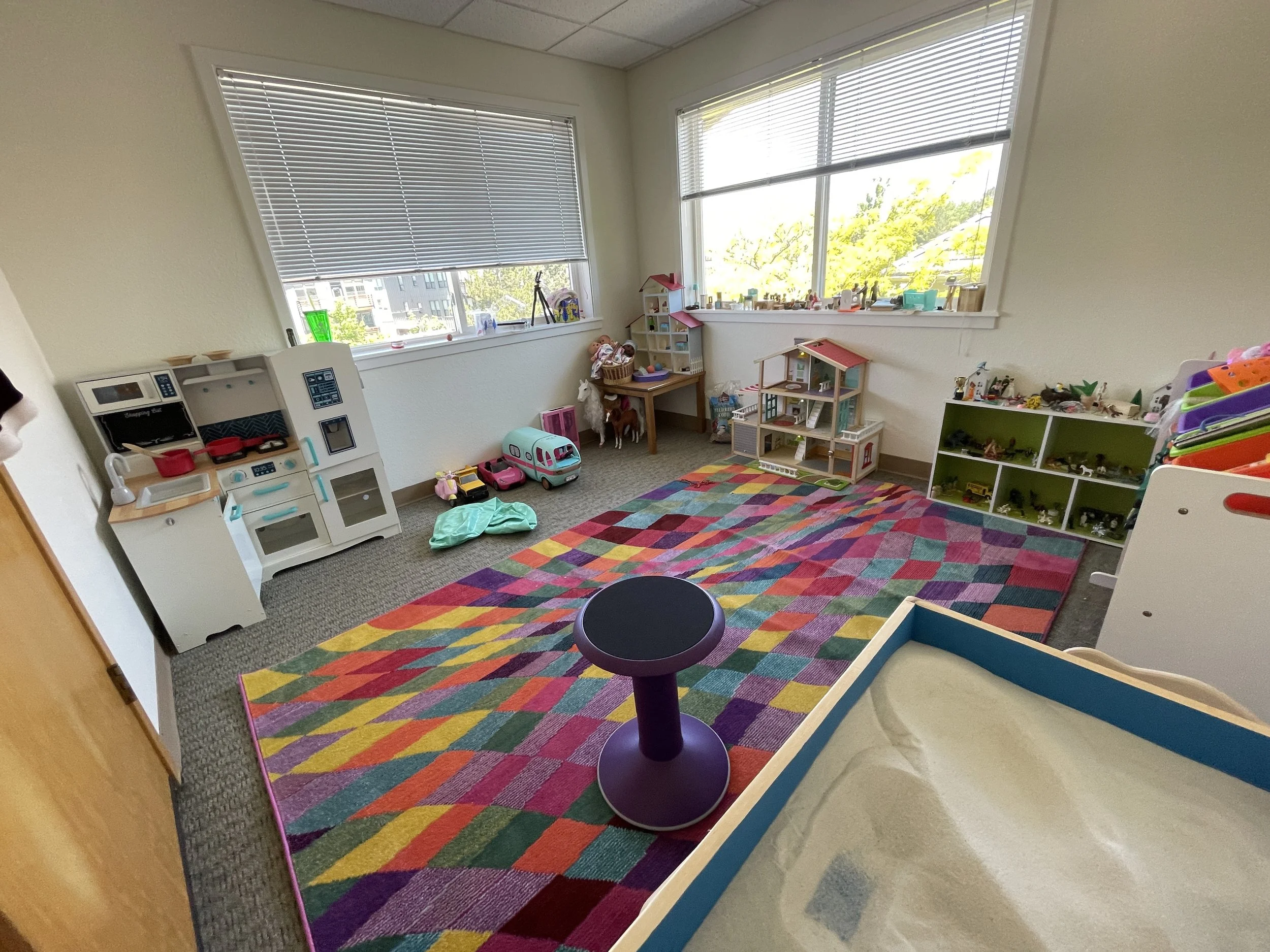 A colorful children's playroom with a multicolored geometric rug, toy kitchen set, playhouse, various toys, and stuffed animals near windows letting in natural light.