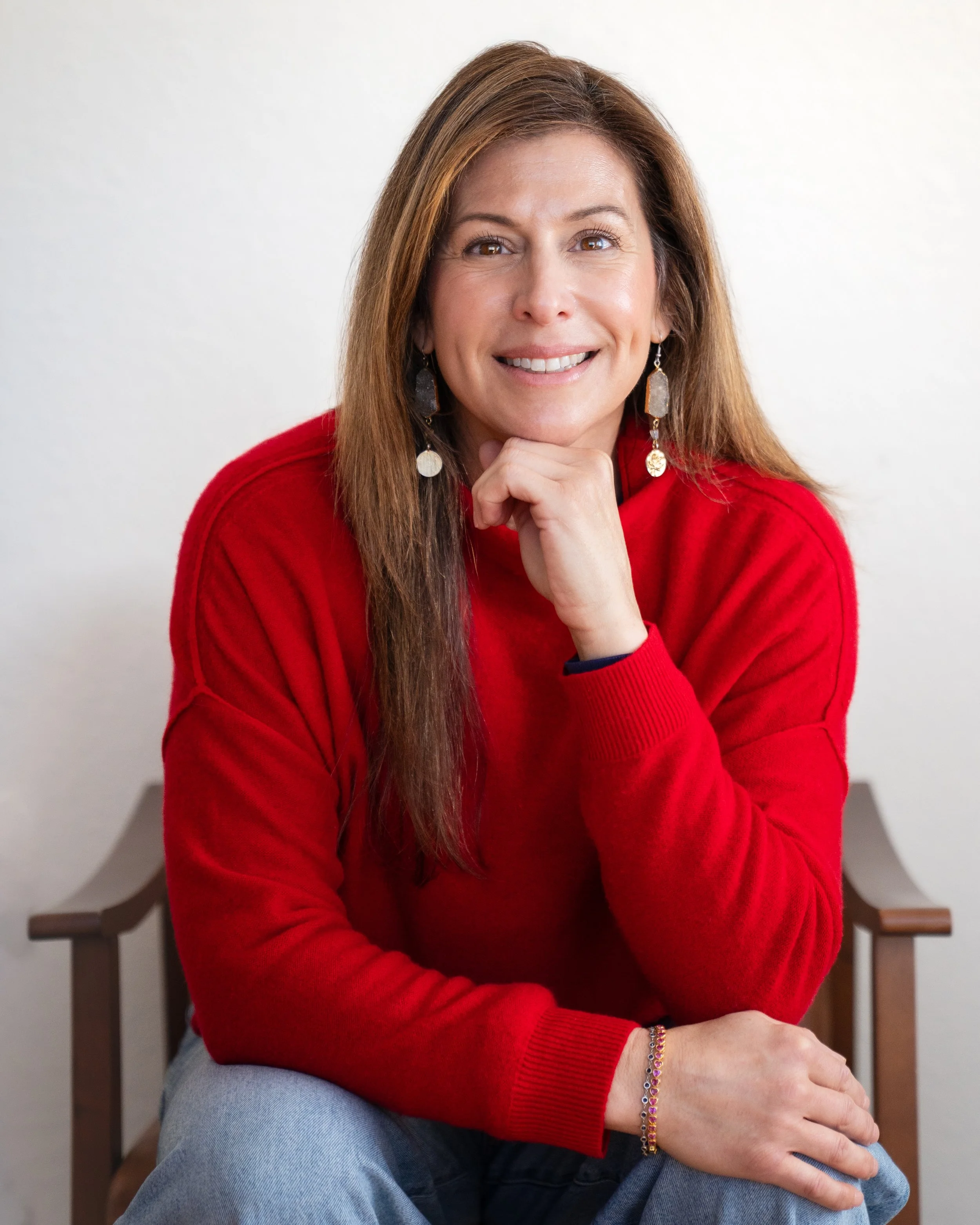 A woman with long brown hair wearing a red sweater, seated on a wooden chair, smiling with her chin resting on her hand, against a plain white wall.