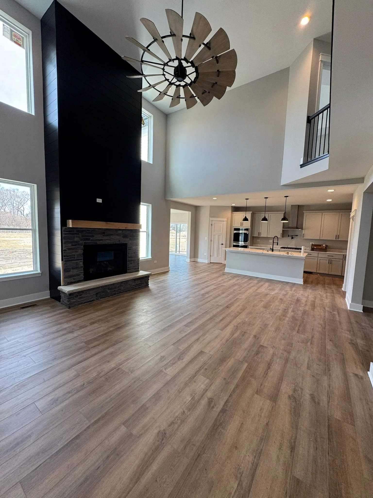Open-concept living room with a high ceiling, a modern black and stone fireplace, large windows, and a view of a kitchen with an island and pendant lighting.