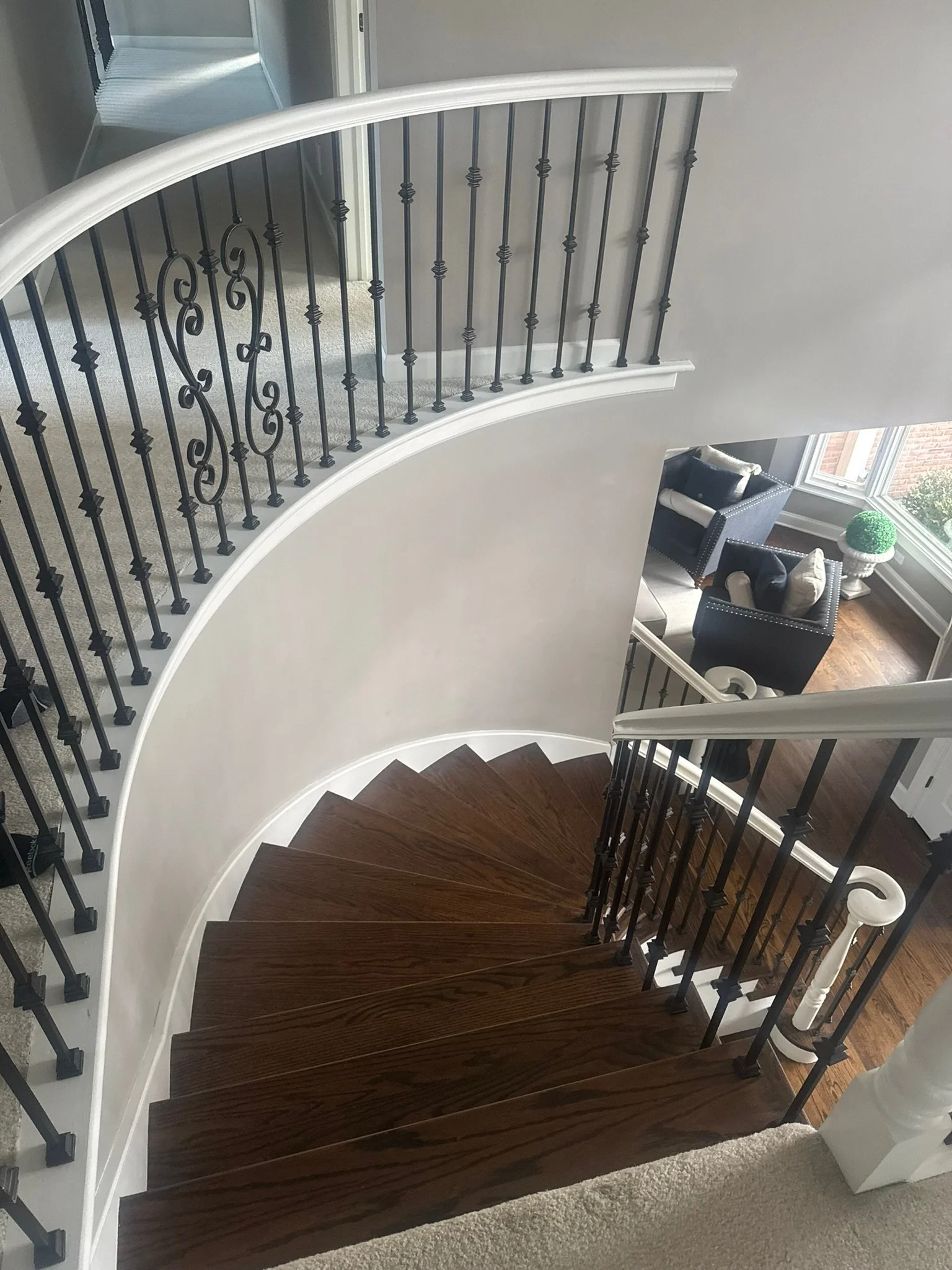 View of a curved wooden staircase with black metal balusters and white handrail, overlooking a living room with black chairs and large windows.