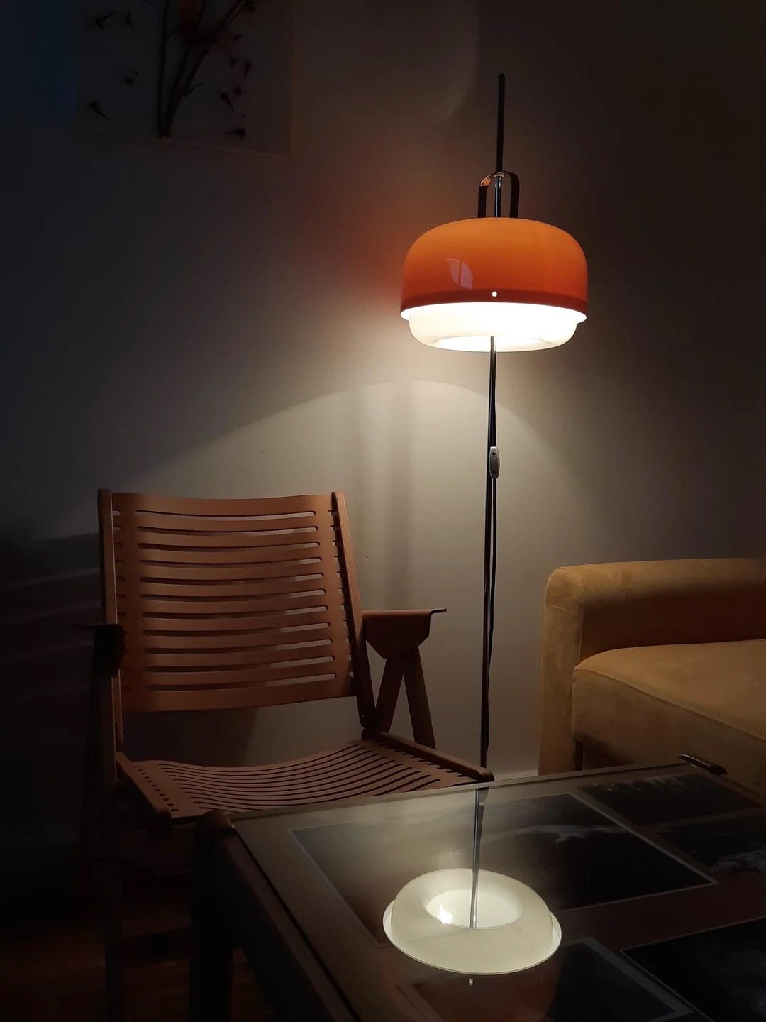 A mid-century modern room with a wooden chair, a yellow sofa, a glass top table, and a floor lamp with an orange and white shade, casting warm light and shadows on the plain wall.