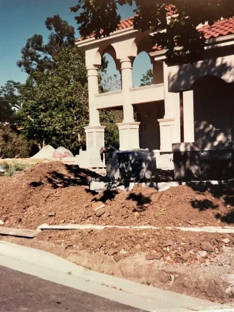 Construction site in front of a building with arched columns and a red tile roof, with dirt and construction tools in the foreground.