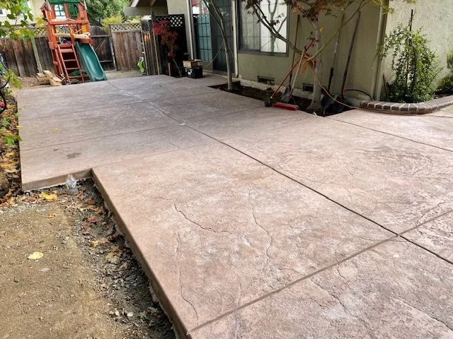 Newly poured concrete patio in backyard with a small step, a shed, a playset, and garden tools in the background, surrounded by a wooden fence and trees.