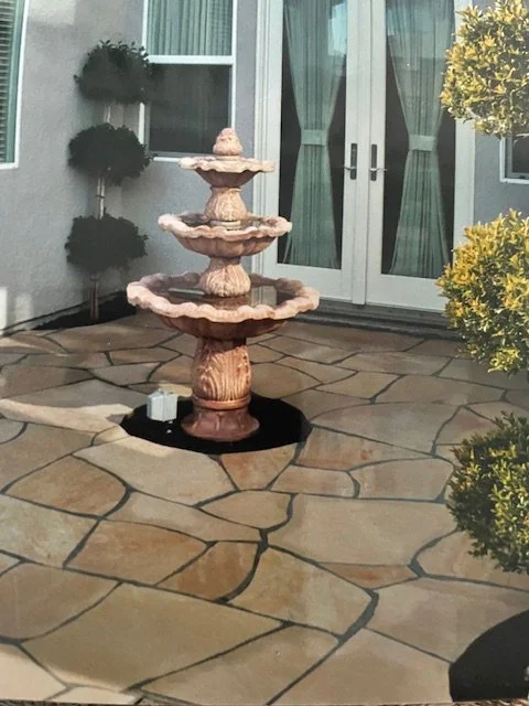 Stone outdoor fountain with three tiers on a paved patio near a house with glass doors and green curtains.