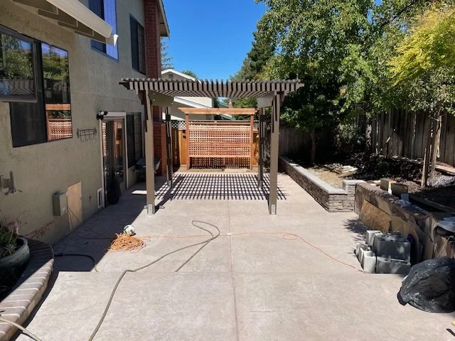 Backyard patio with a pergola, concrete flooring, and a wooden privacy fence, surrounded by trees and a neighboring house to the left.