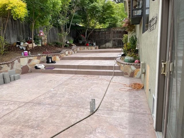 Backyard patio with concrete surface, stone-edged garden beds, trees, and outdoor tools and materials for gardening or construction.