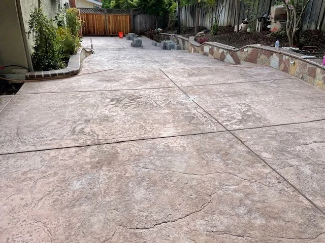 Freshly poured and smoothed concrete patio in a backyard, with some construction materials and tools nearby, bordered by plants and a wooden fence.