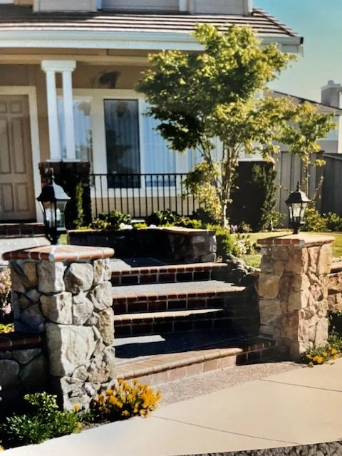 Front yard of a house with stone steps leading to the porch, a small tree, and garden lamps.