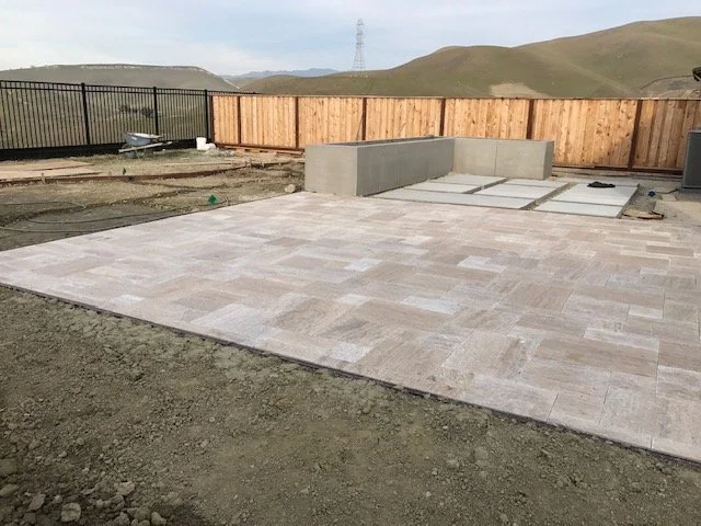 A newly paved outdoor patio area with light-colored tiles, a built-in concrete bench, and a wooden fence in the background, with distant hills and hills in the distance.