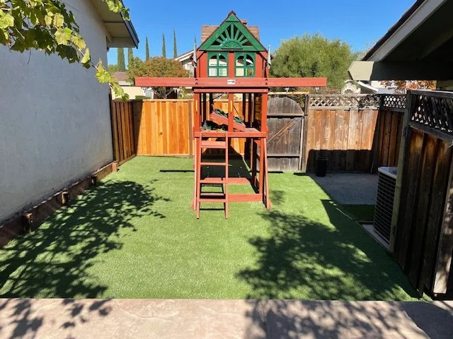 Backyard with a wooden playhouse and slide on artificial grass, enclosed by a wooden fence and shaded by trees.