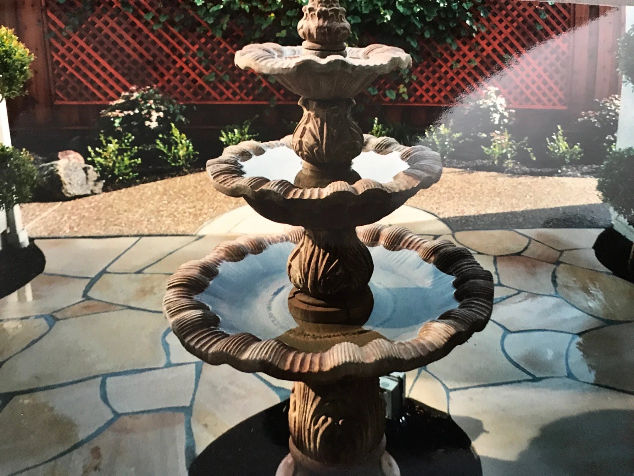 A three-tiered stone fountain with water, surrounded by a paved area, shrubs, and a lattice fence in the background.