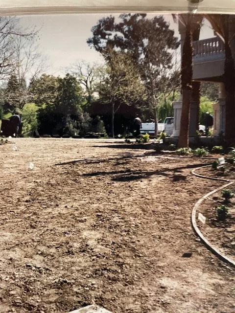 A backyard with a dirt ground, some trees, and construction equipment and vehicles in the background.