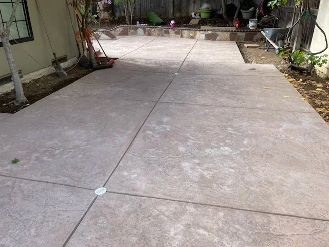 Newly poured concrete patio in a backyard with a drain cover and garden tools to the side.