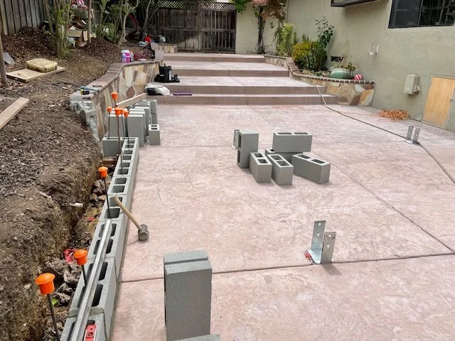 Construction of a new patio or walkway, with concrete blocks and tools in place, and an unfinished section with soil and construction materials.