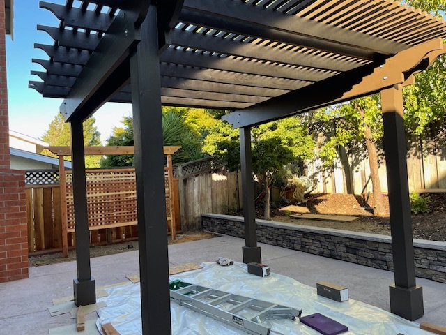 Outdoor patio area under construction with a black pergola, a ladder, and construction tools on a sheet, with a wooden fence, trees, and a brick wall in the background.