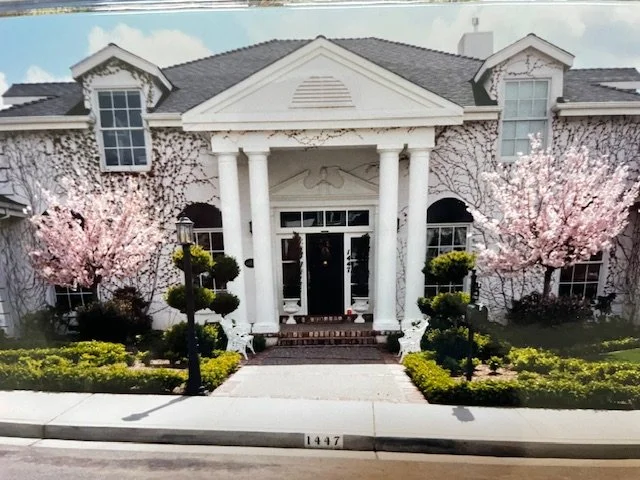 Front view of a large white house with a portico supported by four columns, surrounded by blooming pink trees and trimmed bushes, with house number 1447 on the sidewalk.