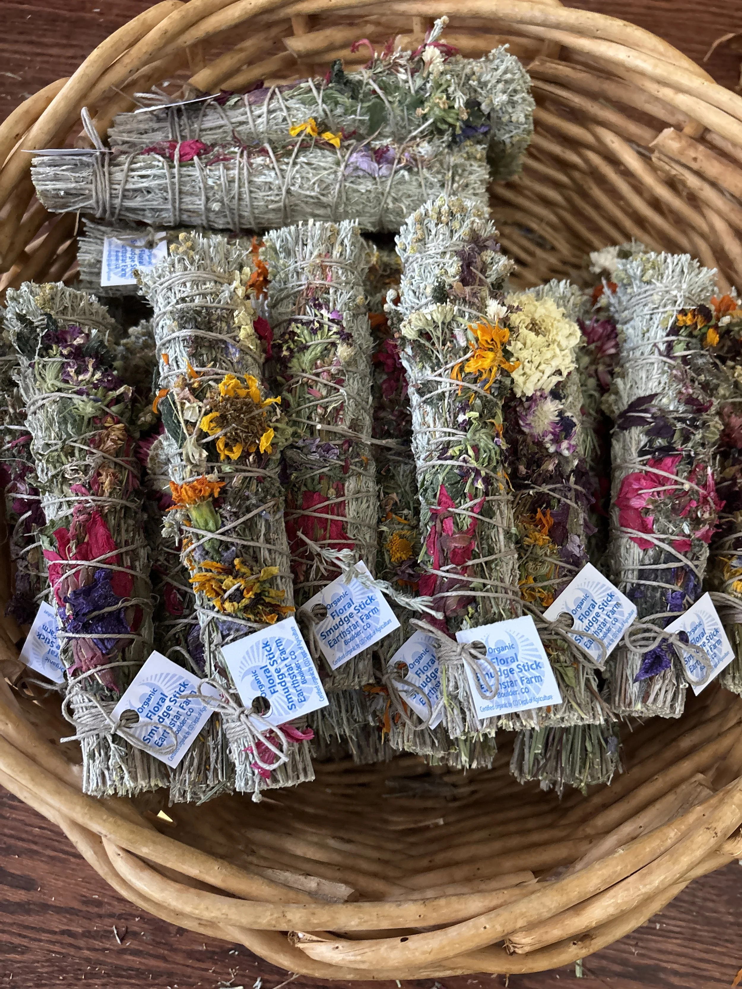 Bundle of dried lavender and other dried flowers tied with string, labeled with tags that read 'Organic Flora Smudge Stick Earthstar Farm,' placed in a wicker basket.