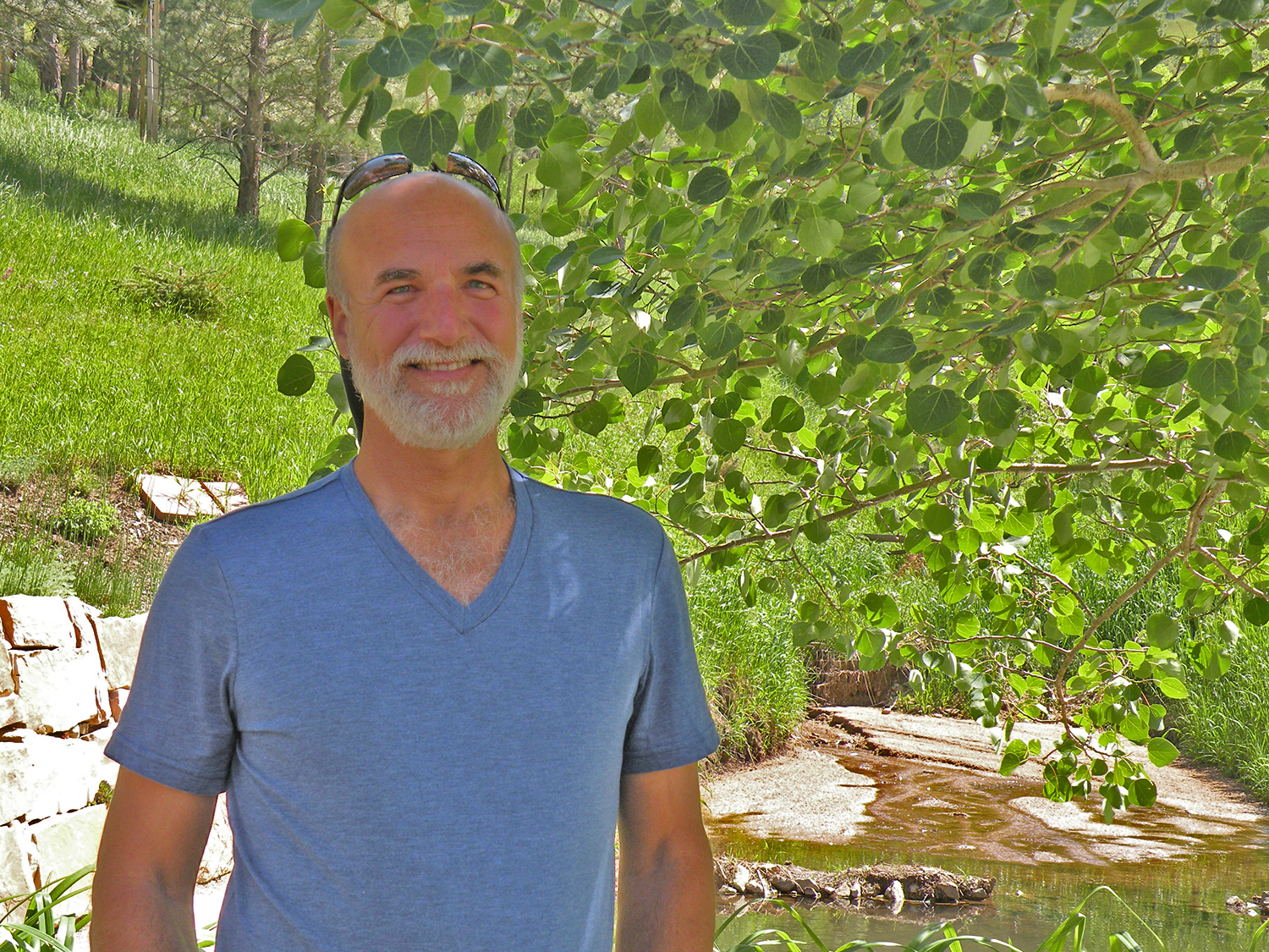 A smiling man with a beard and sunglasses on his head, wearing a blue t-shirt, standing outdoors near a creek with green trees and grass in the background.