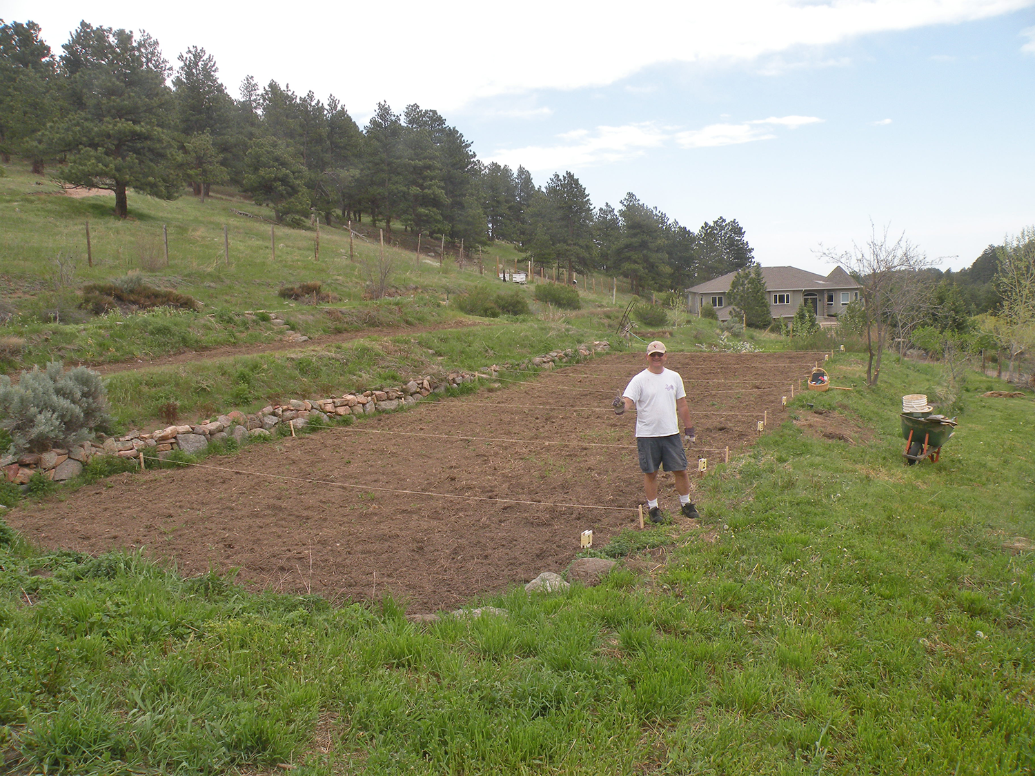 A man stands in a garden plot with tilled soil, holding a gardening glove, surrounded by various gardening tools and supplies on a hillside with a house and trees in the background.