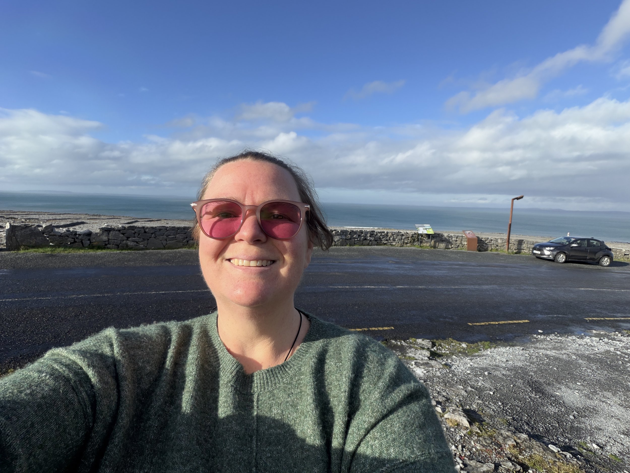 A woman taking a selfie on a coastal road, wearing pink sunglasses and a green sweater, with the ocean and cloudy sky in the background.