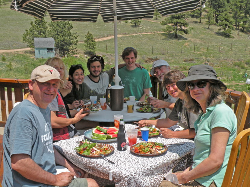 A group of nine people gathered around a table outdoors on a sunny day, enjoying a meal with salads, watermelon, and drinks, under a large striped umbrella with a scenic hilly landscape in the background.