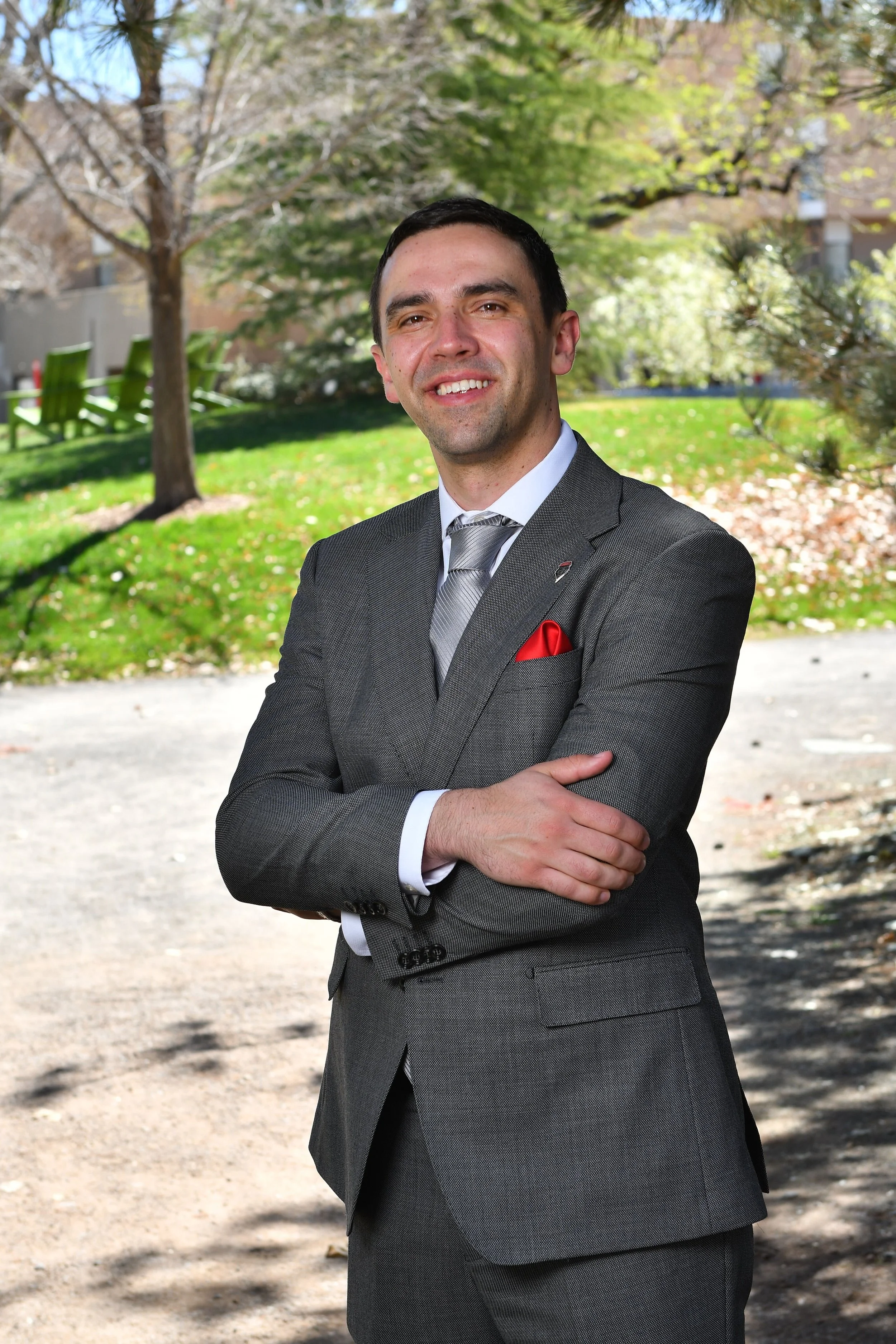 A man in a gray suit with a red pocket square posing with arms crossed outdoors in a park with green grass, trees, and a building in the background.