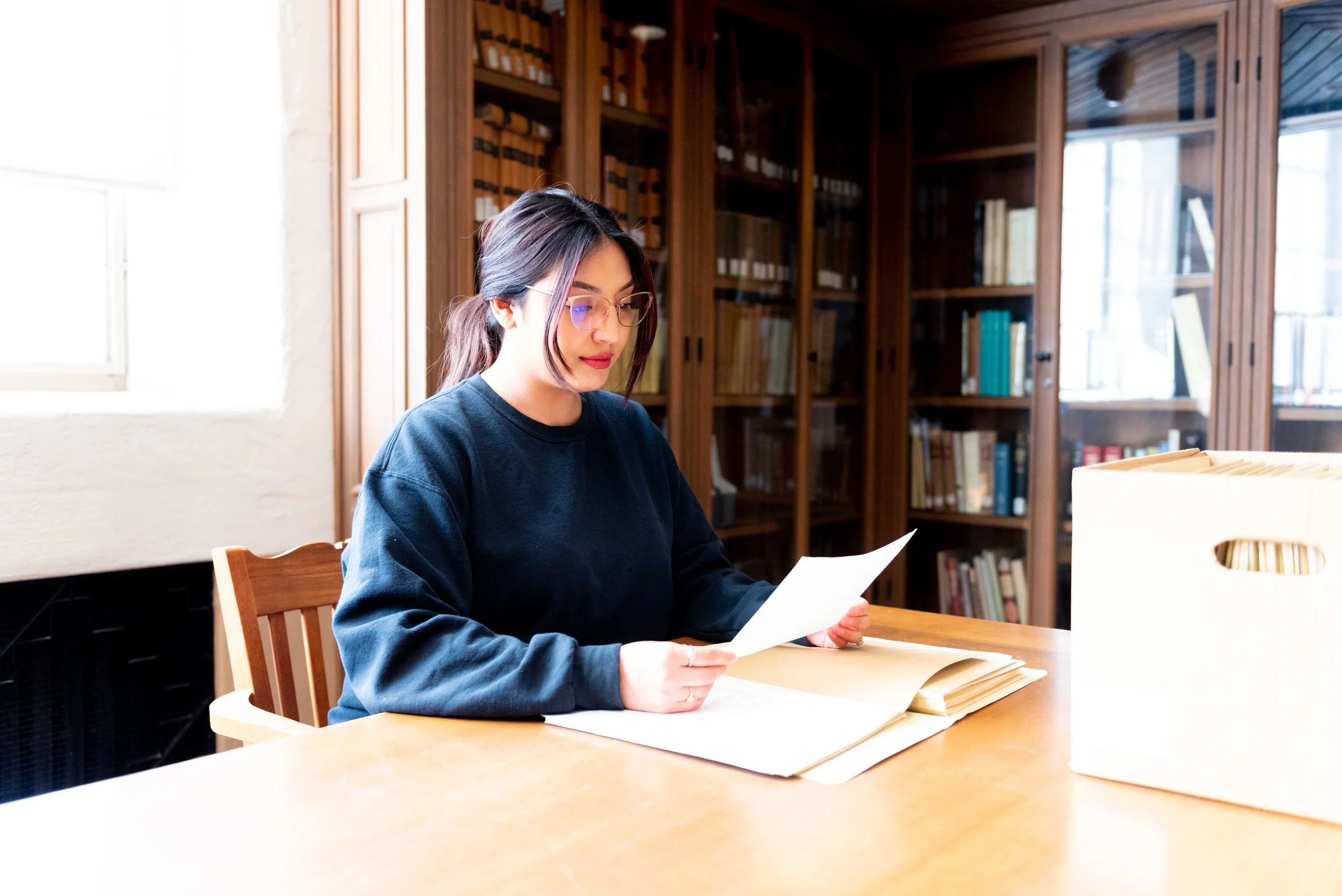 A young woman with glasses and dark hair tied in a loose ponytail sitting at a wooden table in a room with wooden bookshelves. She is reading a document with stacks of papers in front of her.
