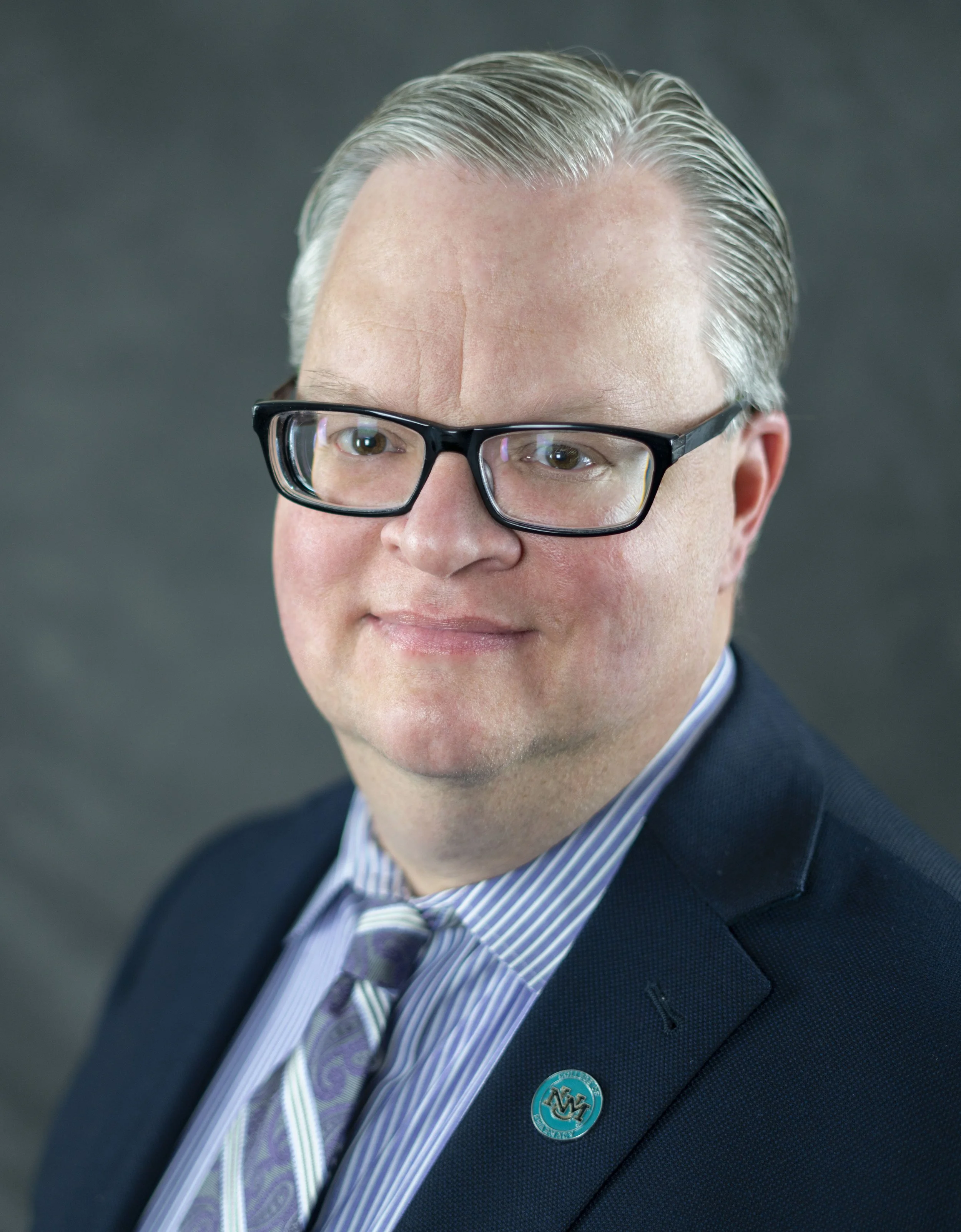 Close-up of a man wearing black glasses, a dark suit, a striped dress shirt, and a patterned tie, with a lapel pin on his suit.