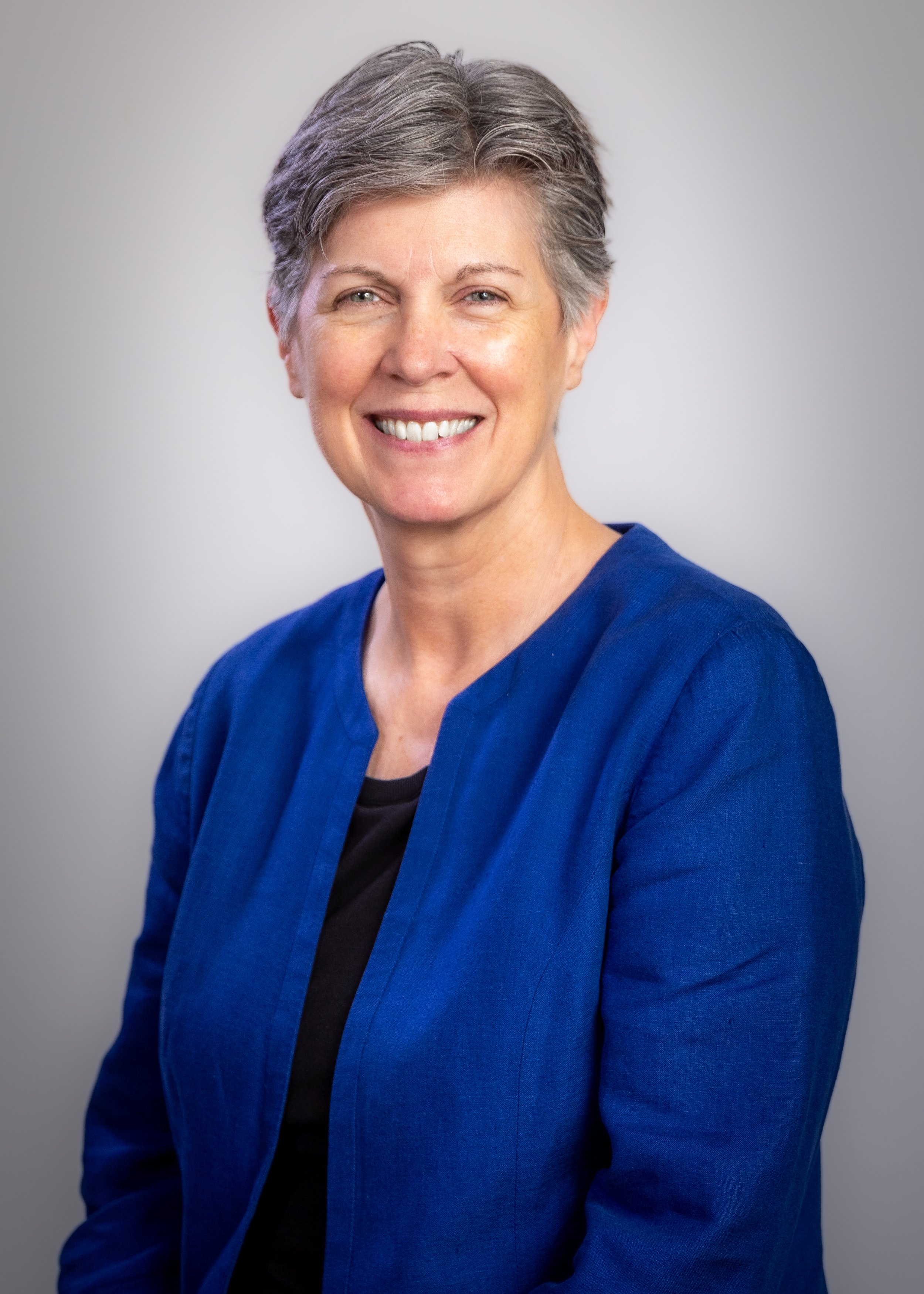 Portrait of an older woman with short gray hair, wearing a blue jacket and black top, smiling against a plain light gray background.