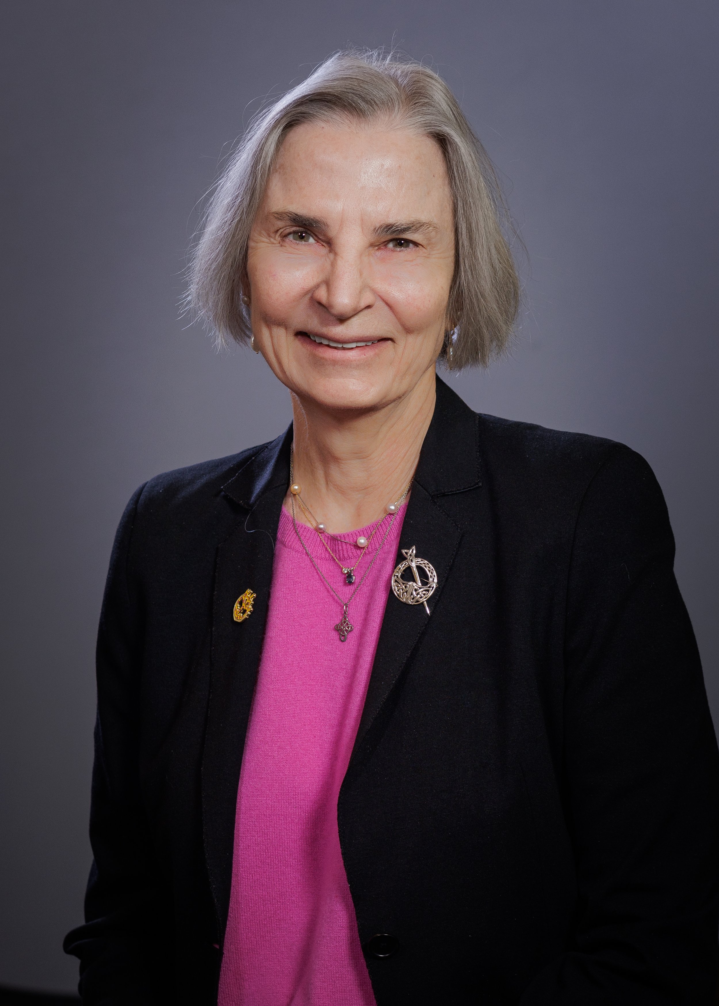 A woman with short, gray hair wearing a black blazer over a pink top, accessorized with jewelry including a pearl necklace and various pins and brooches, smiling against a plain gray background.