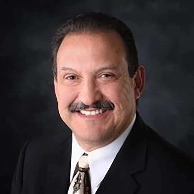A smiling man with a mustache and short dark hair, wearing a black suit, white shirt, and a patterned tie, against a dark background.