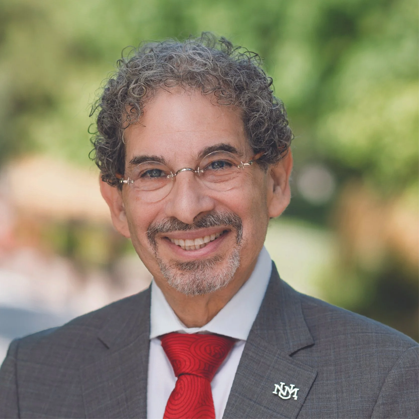 Close-up of a smiling man wearing glasses, a dark suit, a white shirt, and a red tie, with a blurred green outdoor background.